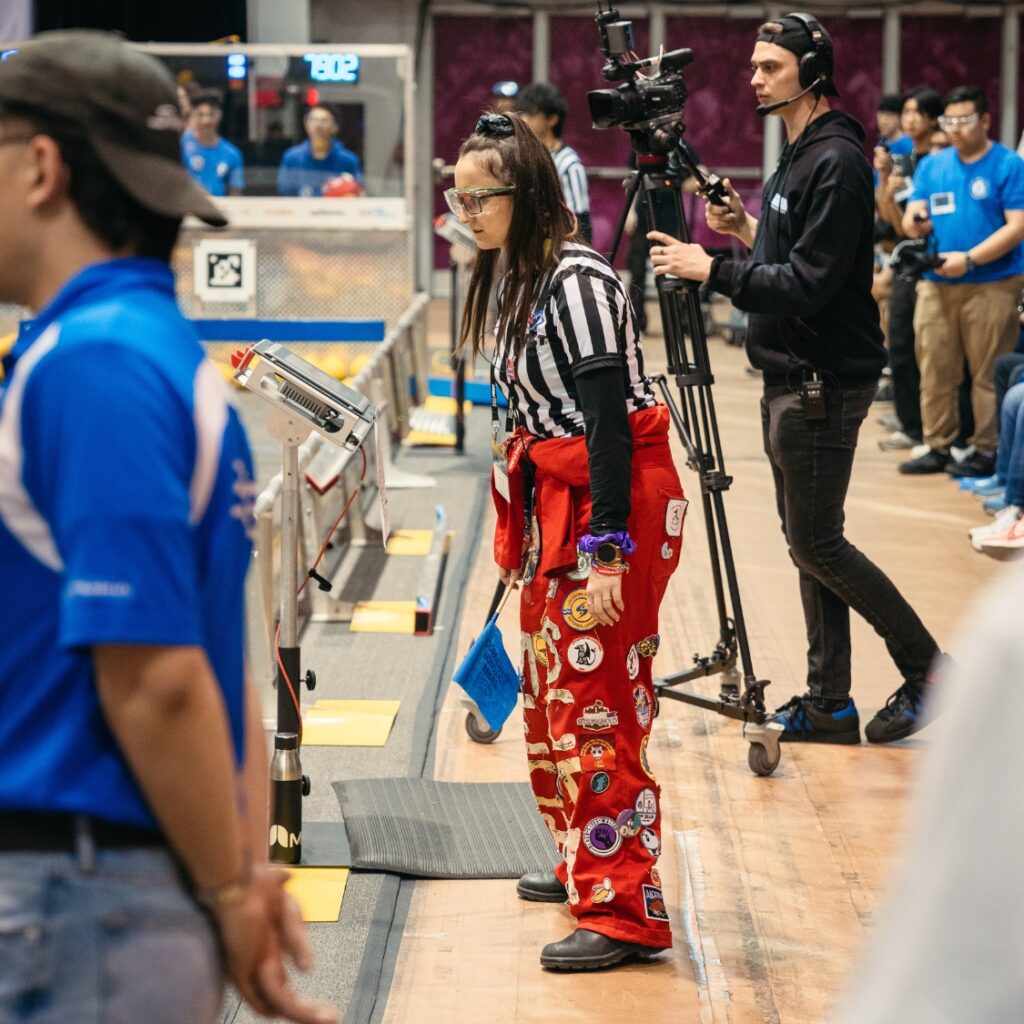 Angela Tollis wearing a red suit and striped ref shirt on the sidelines of the FIRST field