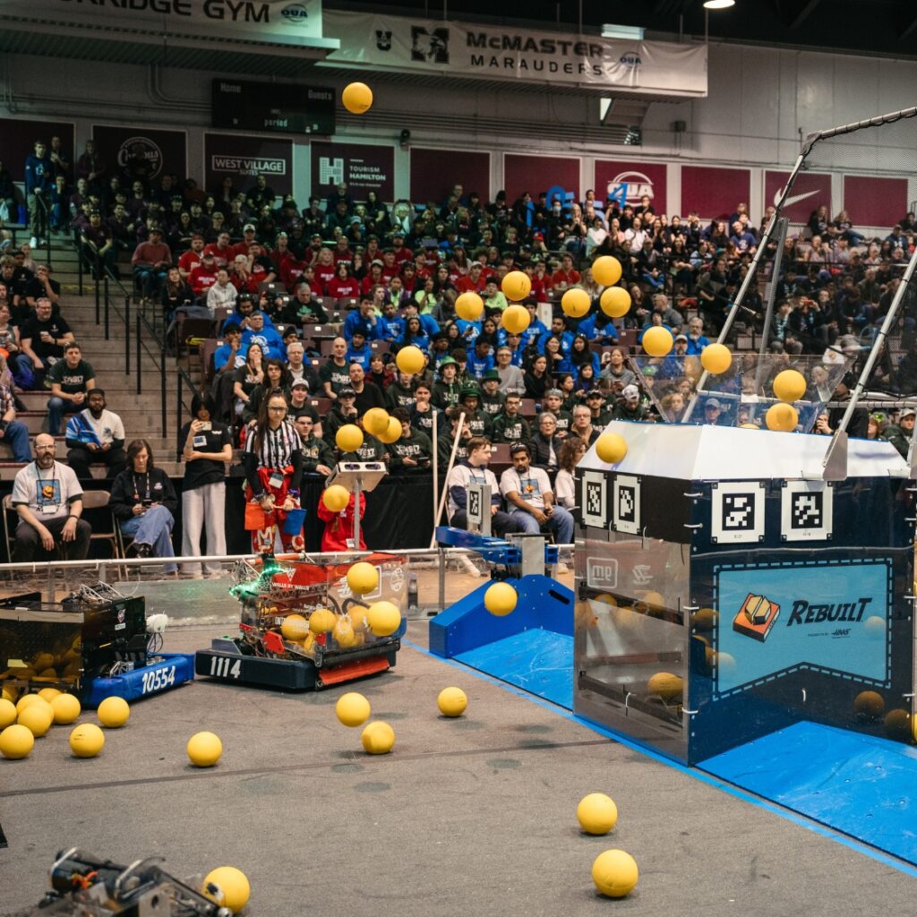 two robots on the field, a slew of balls fly in the air towards a large container in the middle of the field