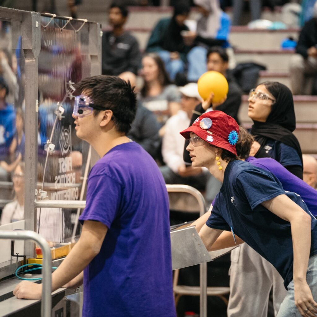 teams on the edge of the field looking on to the action, one person is prepped to shoot a ball.