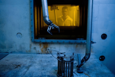 Someone wearing a lab coat stands behind glass, while a robotic arm moves to pick up an object in the foreground.