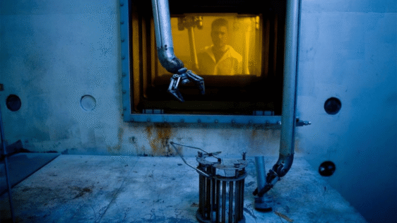 Someone wearing a lab coat stands behind glass, while a robotic arm moves to pick up an object in the foreground.