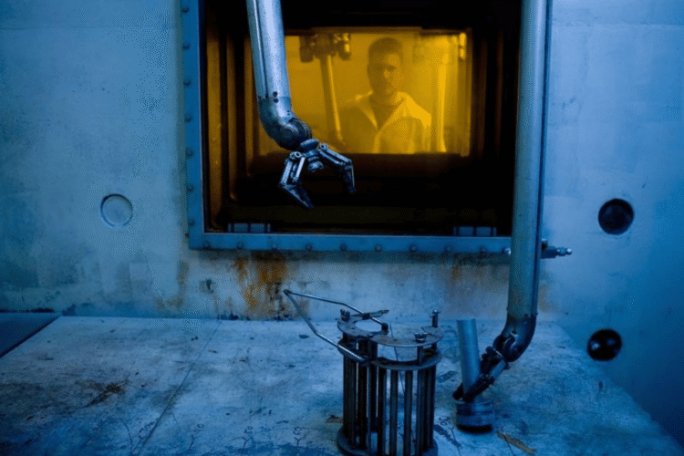 Someone wearing a lab coat stands behind glass, while a robotic arm moves to pick up an object in the foreground.
