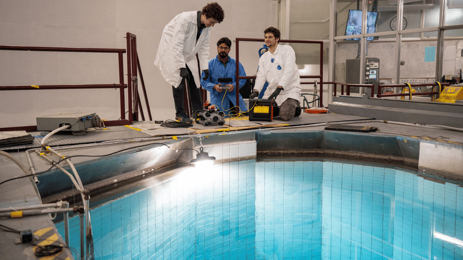 Three Engineering Physics students wearing lab coats prepare an underwater drone by the water of the McMaster Nuclear Reactor.