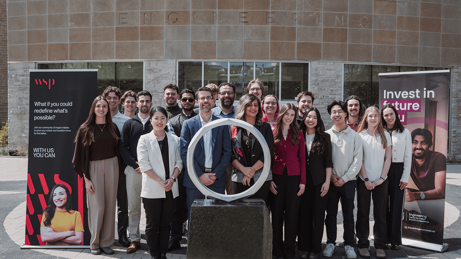 A group of people stand behind an iron ring statue outside of JHE with WSP and McMaster Engineering branded banners.