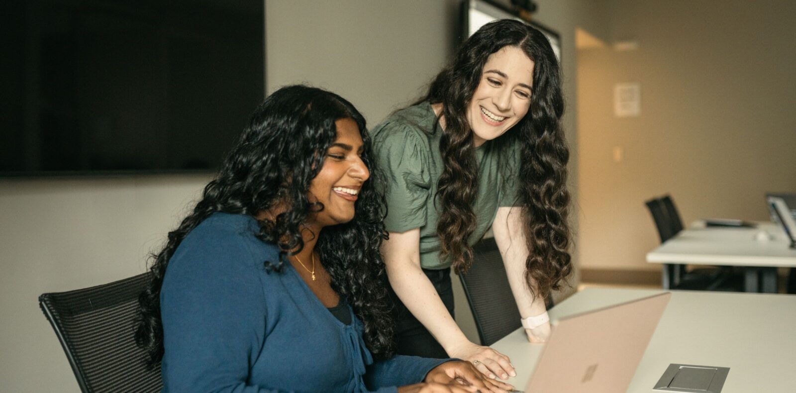 A student works on a laptop and smiles while her supervisor oversees her work