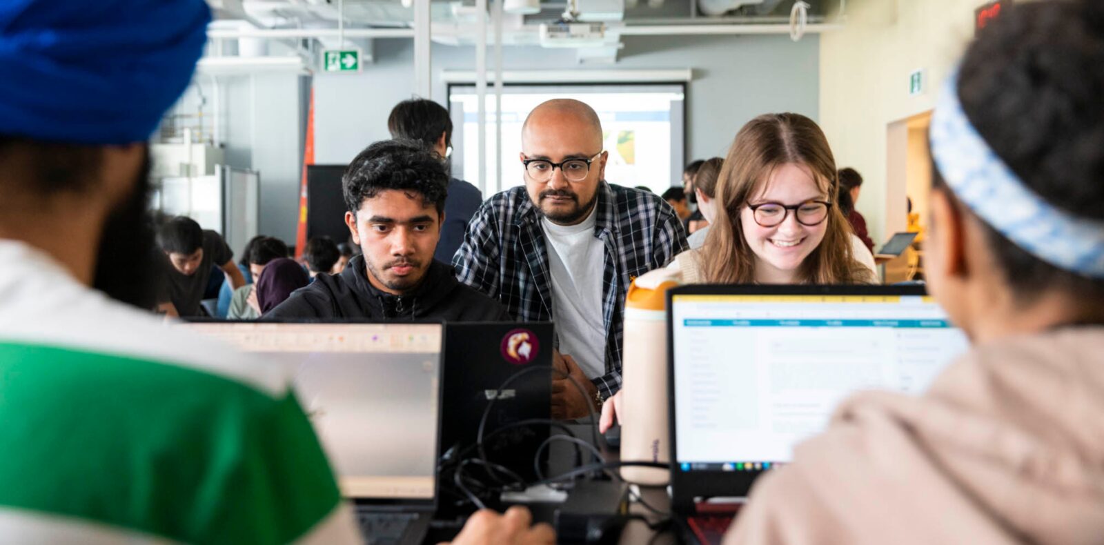 Students sitting at a table in a design studio working on an engineering project while an instructor observes their progress