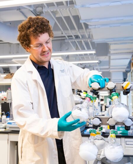 A person wearing a lab coat holds a round-bottomed flask up to a frame.