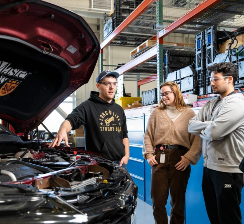 Someone gestures to parts under the hood of a car while two other people stand to the side.