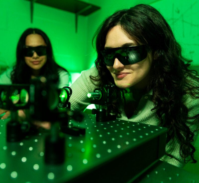 Two people wearing dark safety glasses work with machinery featuring a green beam.
