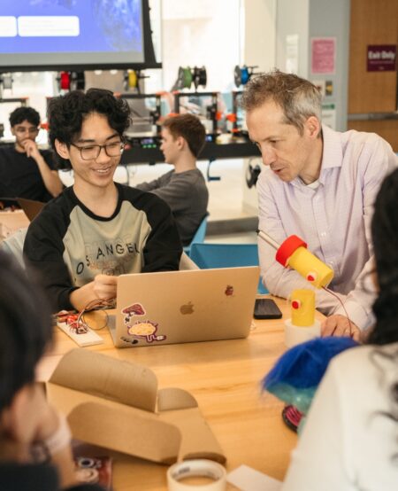 A professor leans over 3D printed components while a student works on a laptop.