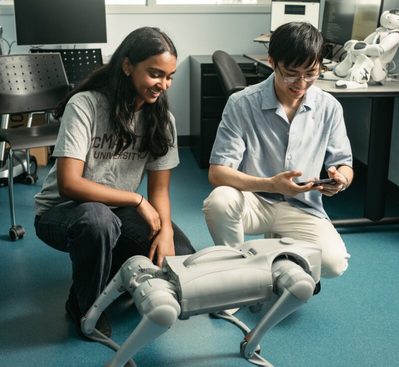 Two people, one holding a controller, kneel beside a robotic dog.