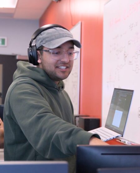 A student wearing headphones holds a laptop and gestures to another computer screen.