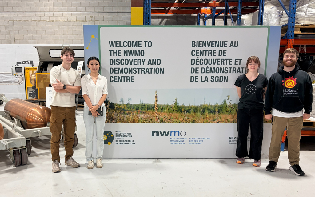 McMaster students Aren, Yuri, Camille and Cole pose in front of a sign at the Nuclear Waste Management Organization. The sign reads Welcome to the NWMO Discovery and Demonstration Centre.