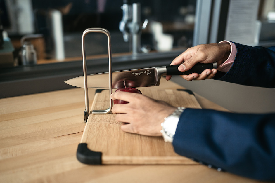 Hands cutting an apple on a cutting board. The blade of the knife is inside an assistive cutting tool that has been slipped onto the side of the cutting board. 